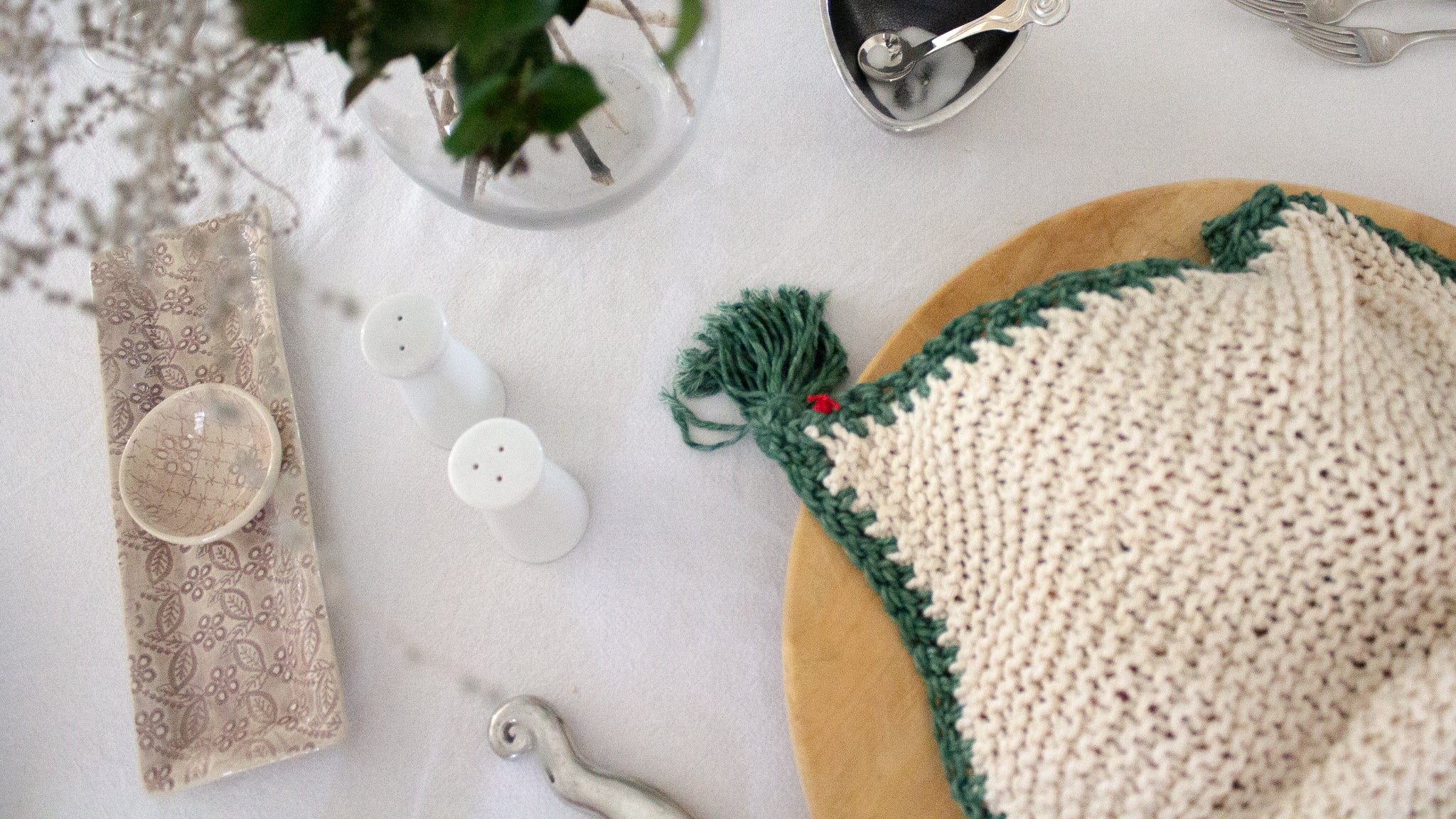 Birds-eye view of a Penelope D Judaica, Duo Colour Tassel Challah Cover in Ivory with Sage Green edging and tassels, covering challah on a round wooden bread board on a set table. 