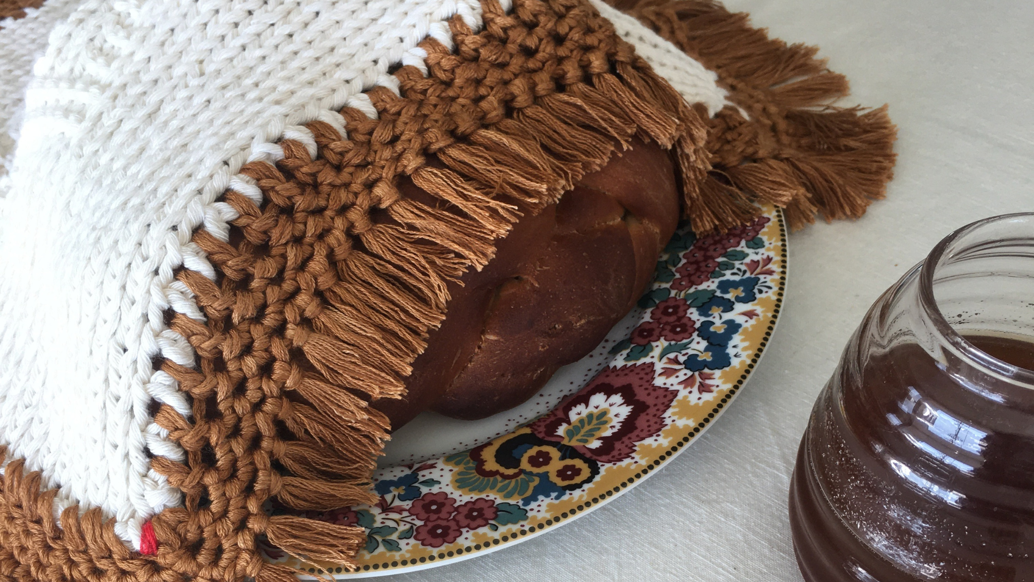 A Penelope D Judaica, Fringes Challah Cover, in ivory and copper edging on a plate with a honey jar.