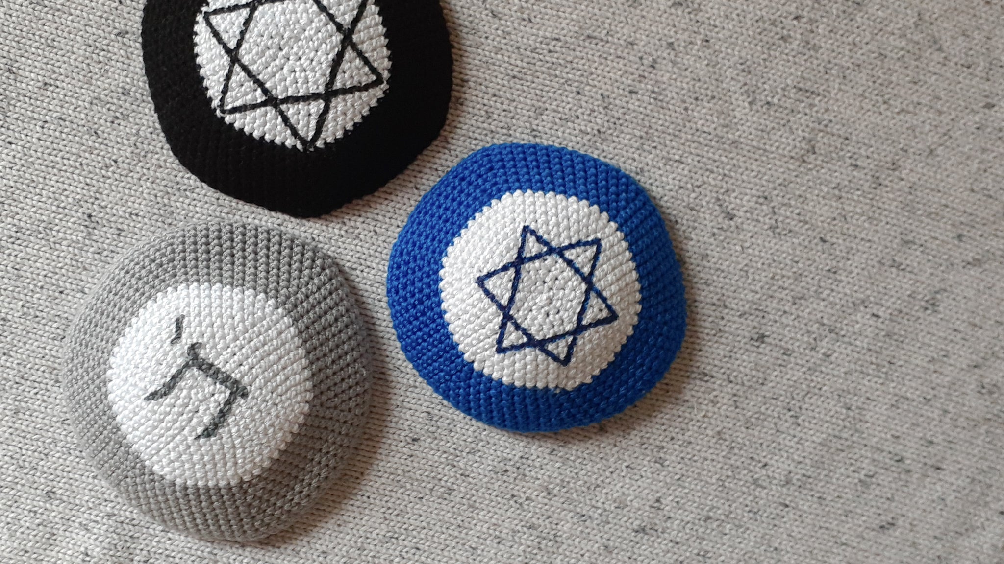 Penelope D Judaica, 3 Jewish Symbol Kippot on a grey knit flatlay. Black & white with a large Magen David, blue & white with a standard Magen David & grey and white Chai colour combination.