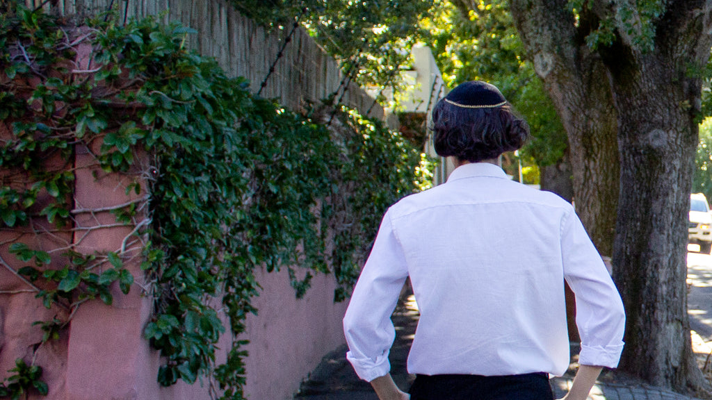 Male walking outside, past a pink and ivy covered wall, dress smartly in a white shirt and black pants, wearing his gold edged black Penelope D Judaica Metallic Edged Kippah.