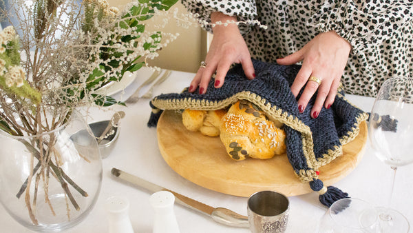 Woman in a floral black and white dress, with her hands over 2 challahs on a round wooden board, covered with a Penelope D Judaica, Metallic Edged Tassel Challah Cover, in charcoal and gold edging, on a set and decorated table.